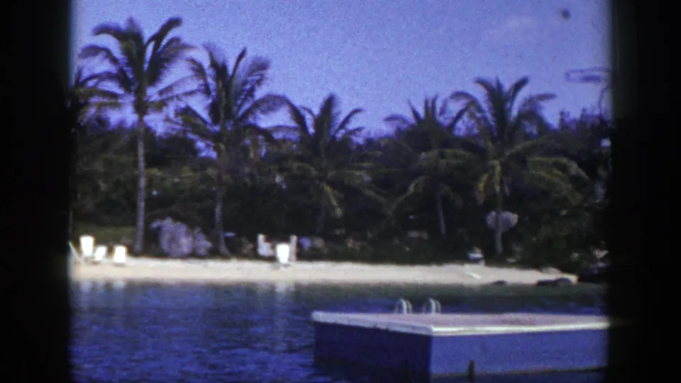 1948 home-movie frame showing palms and lounge chairs along a beach, with blue water and the edge of a floating dock in front.
