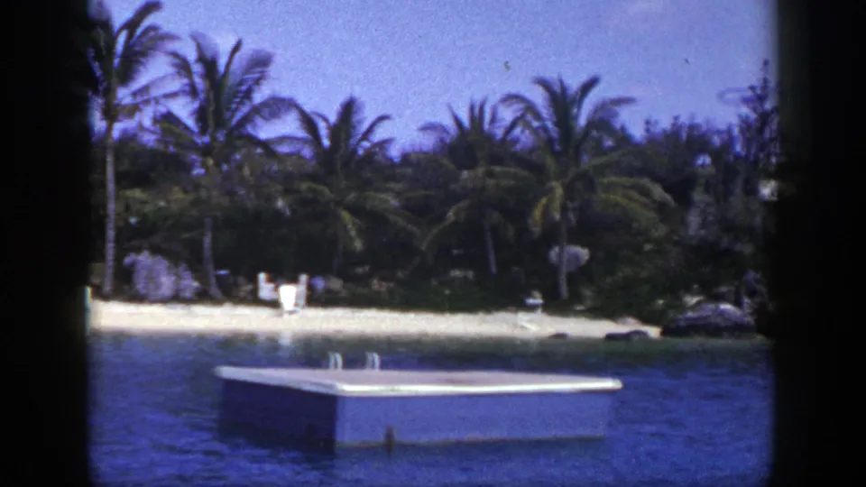 1948 8mm frame showing a small floating dock in the foreground with palms, beach chairs, and a sandy shoreline behind it.