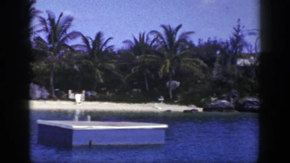 1948 8mm home-movie frame of a palm-lined beach seen across blue water, with a small floating dock and distant figures near the sand.