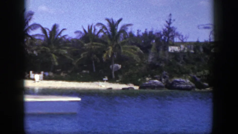1948 8mm frame looking toward a palm-lined shore with blue water, pale sand, rocks, and distant people near the beach.
