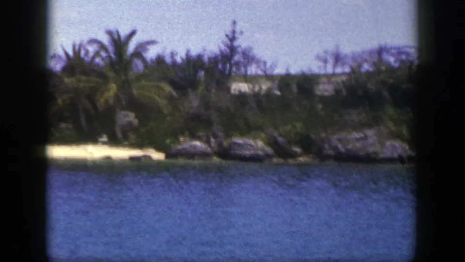 1948 8mm frame of blue water and a narrow palm-lined beach, with rocks and shoreline vegetation visible beyond the sand.