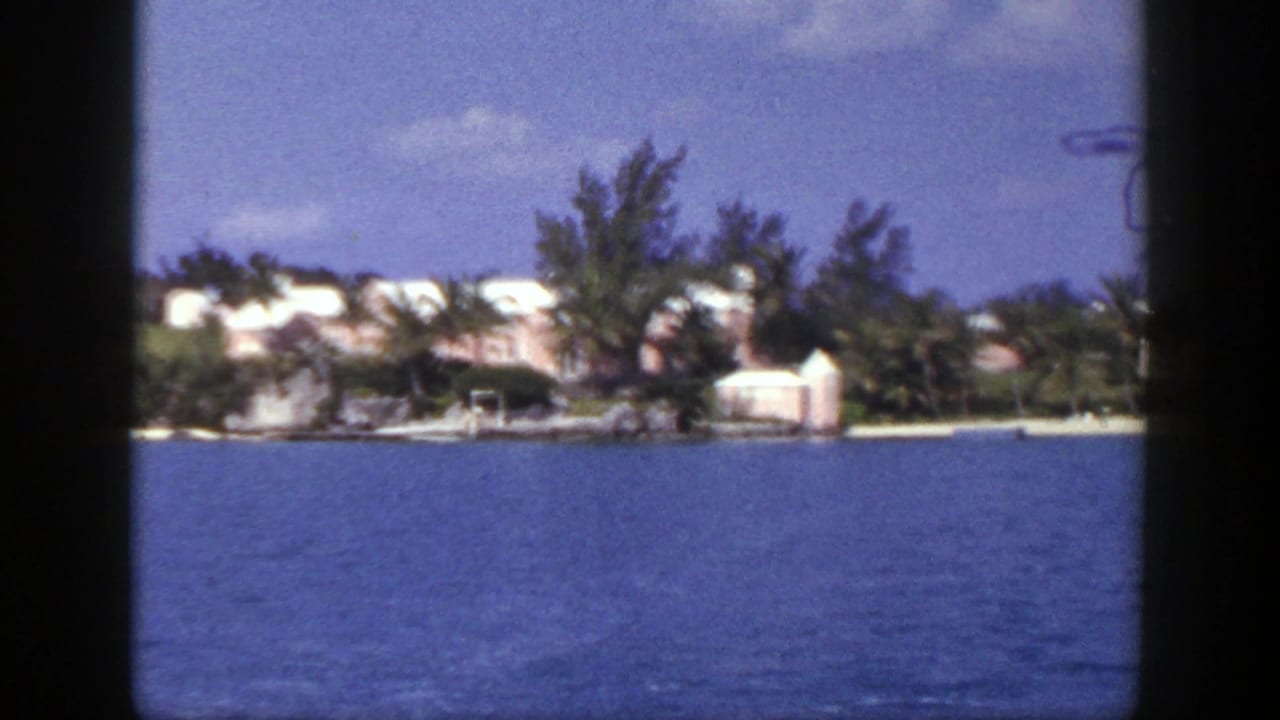 1948 New York-labeled 8mm frame showing blue sea, a pale coastal mansion, palms, and shoreline trees from across the water.