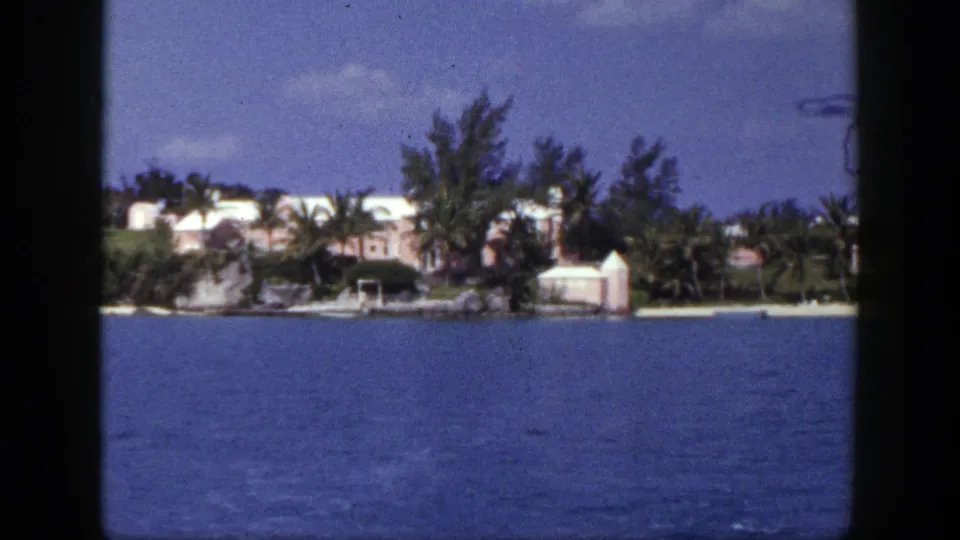1948 New York-labeled home-movie frame with a pale seaside residence, palms, trees, and calm blue water viewed from offshore.