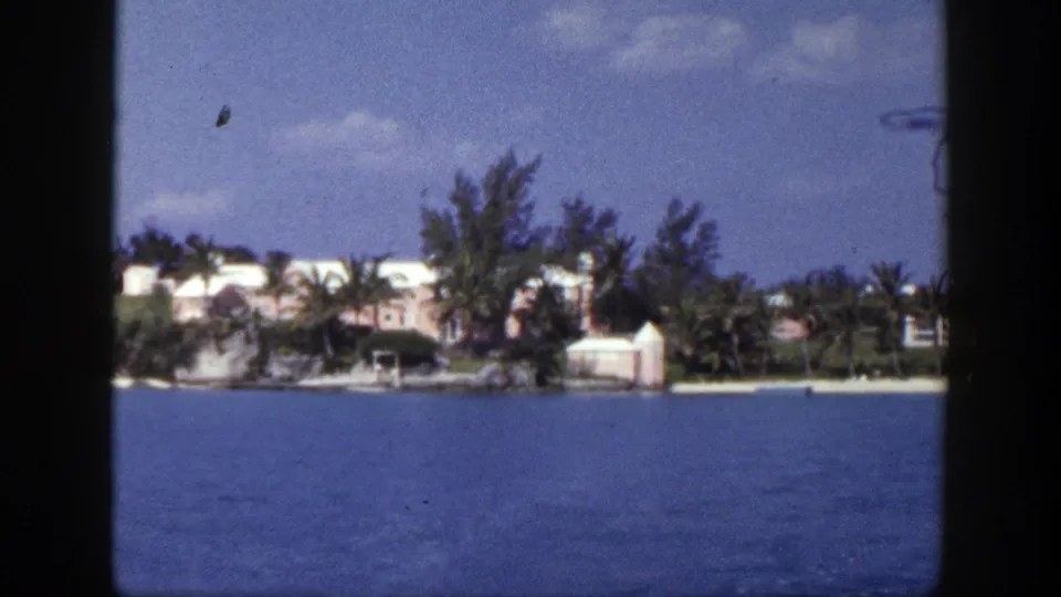 1948 New York-labeled 8mm frame showing a distant light-colored waterfront mansion, palms, shoreline trees, and blue sea.