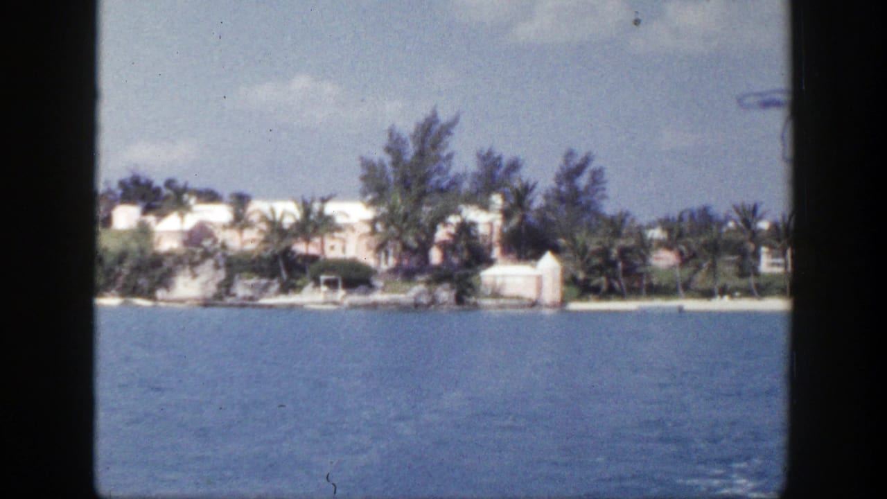 1948 8mm frame showing calm water from a vessel, with a distant shoreline, white buildings, trees, and light clouds.