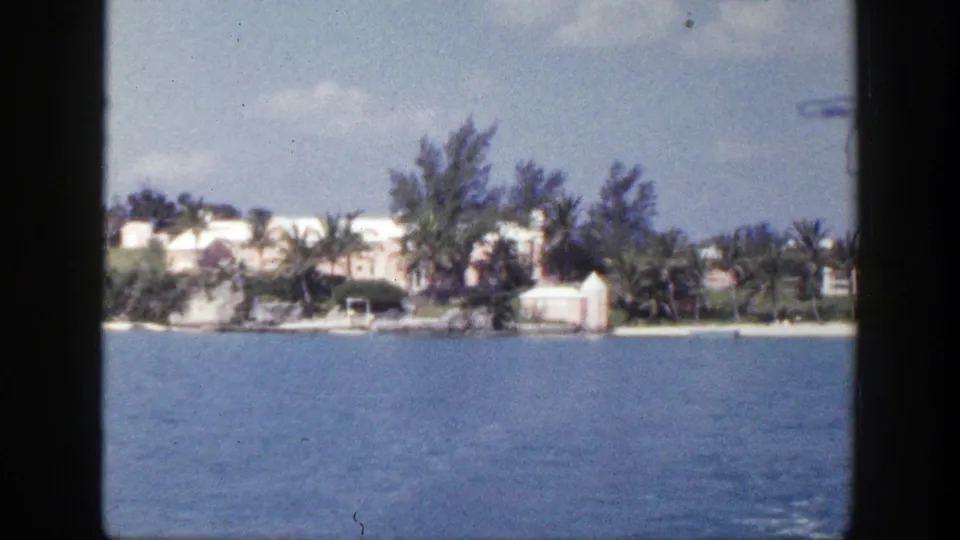 1948 8mm frame showing calm water from a vessel, with a distant shoreline, white buildings, trees, and light clouds.