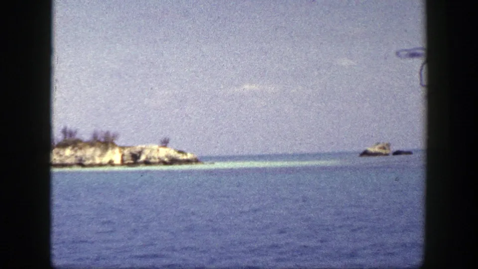 1948 8mm frame looking across blue water toward low rocky islets and a pale sky, with the vessel viewpoint implied by framing.