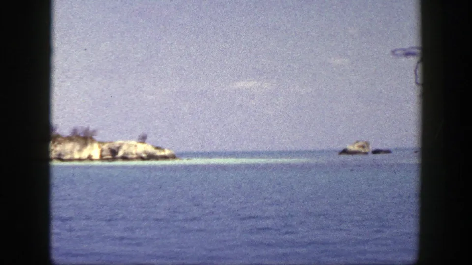 1948 8mm frame of a low rocky islet and small offshore rocks seen across blue water under a hazy sky.