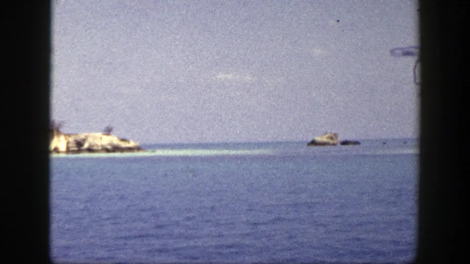 1948 8mm frame showing a rocky islet, small offshore rocks, open blue water, and pale sky within dark film borders.