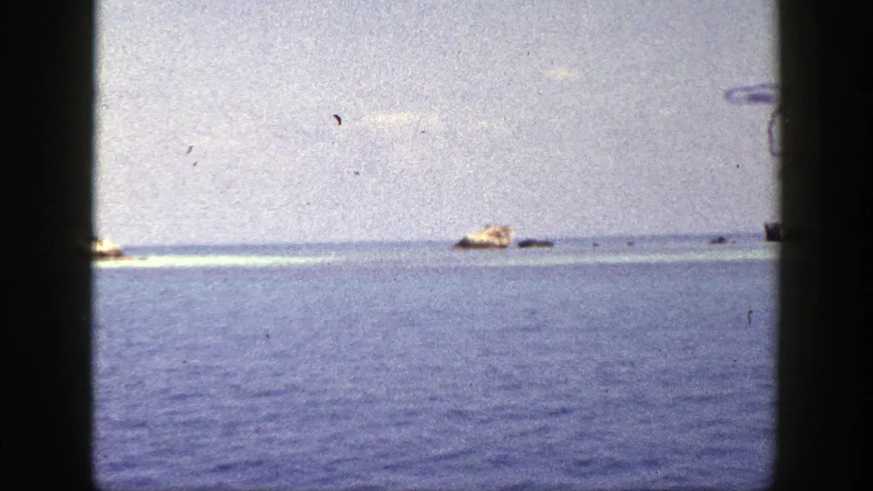 1948 New York coastal frame showing calm ocean, distant tan rocks, and dark window-like borders from a vessel viewpoint.
