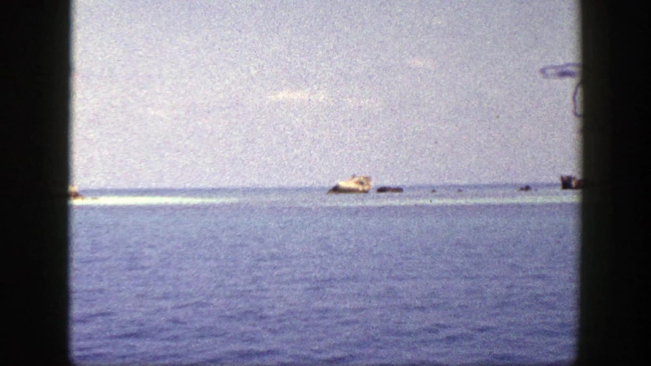 1948 New York 8mm frame showing calm blue water, a pale sky, and small distant tan and dark shapes near the horizon.