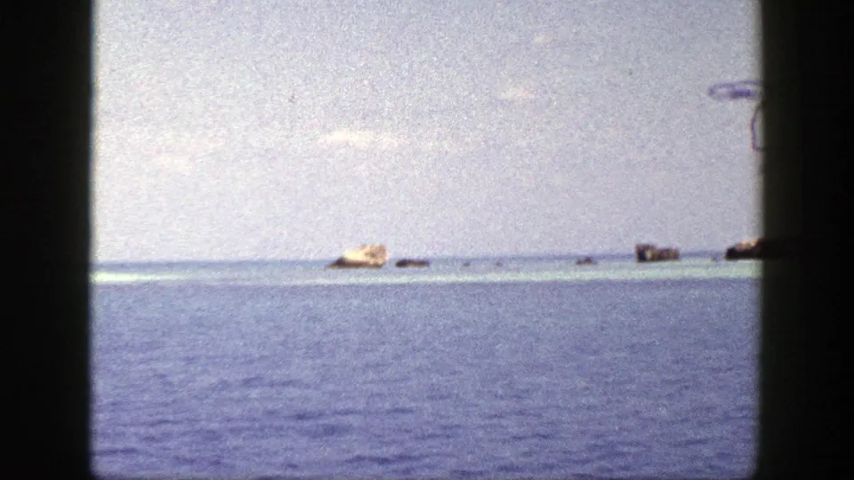 1948 New York 8mm frame showing calm blue water, a pale sky, and distant tan and dark objects near the waterline.