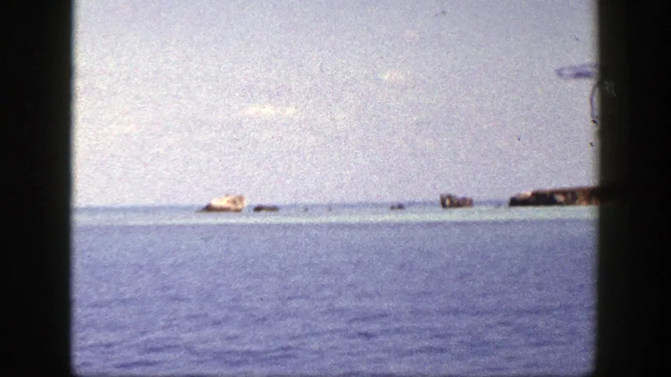 1948 8mm boat perspective showing calm water, a pale distant island or sandbar, and small rocks beneath a washed sky.