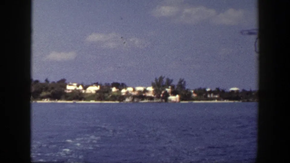 1948 New York home-movie frame looking from a boat across blue water toward a tree-lined shore with white buildings and clouds.