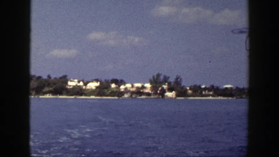 Boat-facing view across blue water toward a distant New York shoreline with pale buildings, trees, and scattered clouds.