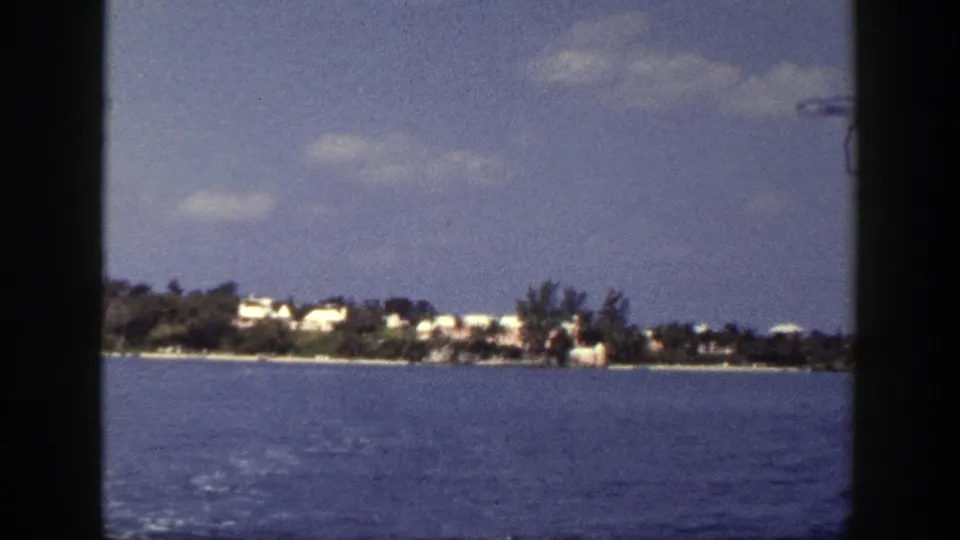 Soft 1948 8mm frame showing rippled blue water and a low tree-lined shoreline with small white buildings in the distance.