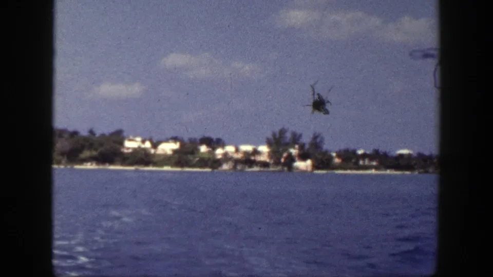 New York coastal home-movie frame with blue water, distant white buildings, trees, clouds, and a visible dark film mark near the sky.