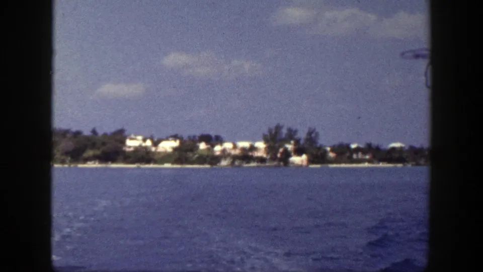 Soft 8mm frame from 1948 New York showing open water, a low treed shoreline, pale buildings, and a clouded sky.