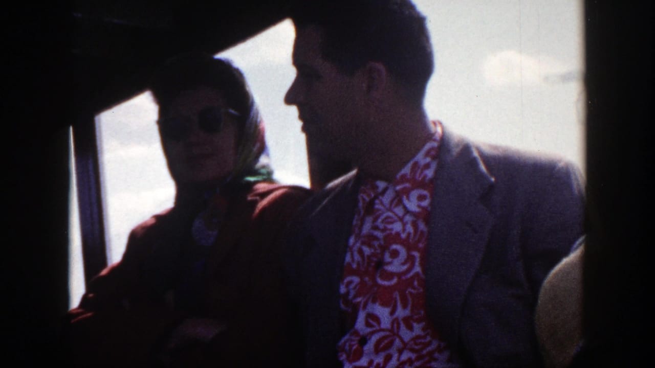 Two adults seated inside a bus, with a woman in sunglasses beside a man in a suit.