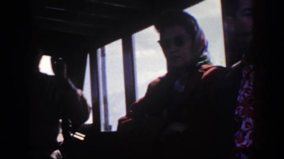 A seated woman wearing sunglasses and a headscarf is framed by a bright bus window, with a suited passenger partly cropped beside her.