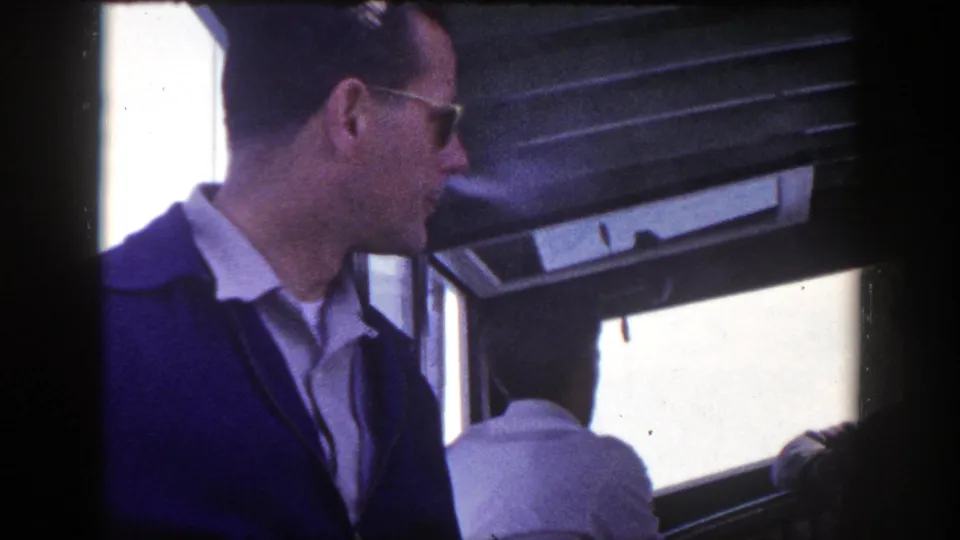 1948 New York 8mm frame showing a man in sunglasses smoking beside seated passengers in a dim travel interior.
