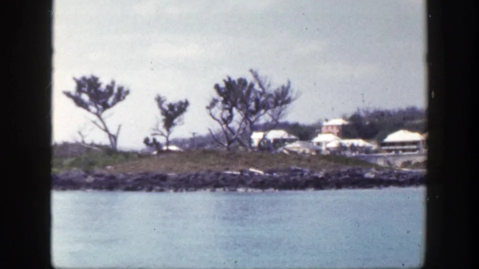 1948 New York 8mm frame of a low coastal shoreline, blue water, sparse trees, and distant tan and white buildings.