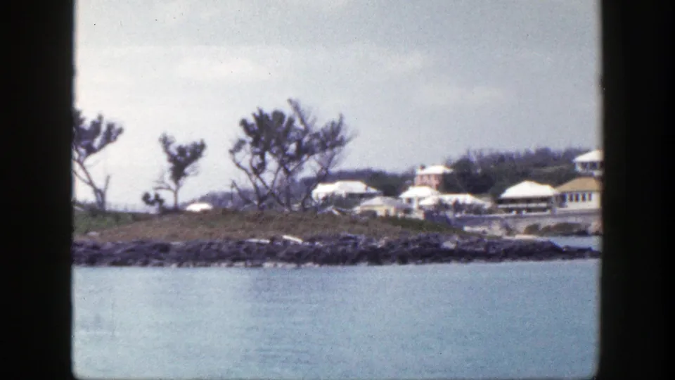 1948 New York 8mm frame showing more of the coastal buildings, rocky shore, sparse trees, and calm blue water.