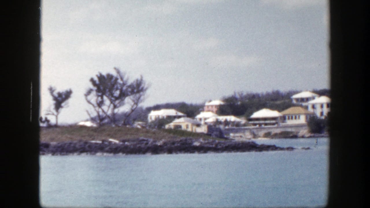 Distant white buildings on a rocky shoreline across calm blue water under a cloudy sky.