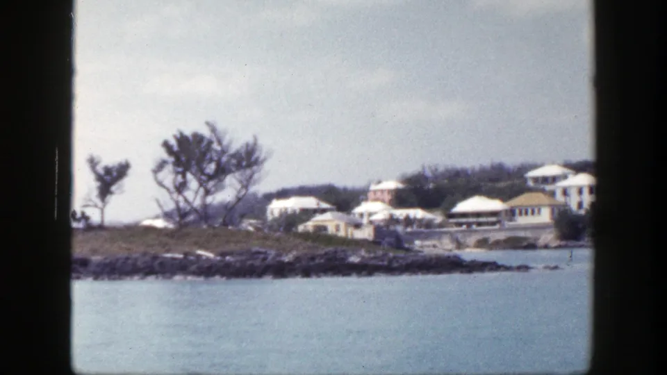 1948 New York home-movie frame showing pale waterfront buildings, trees, rocks, and blue water under soft cloud cover.