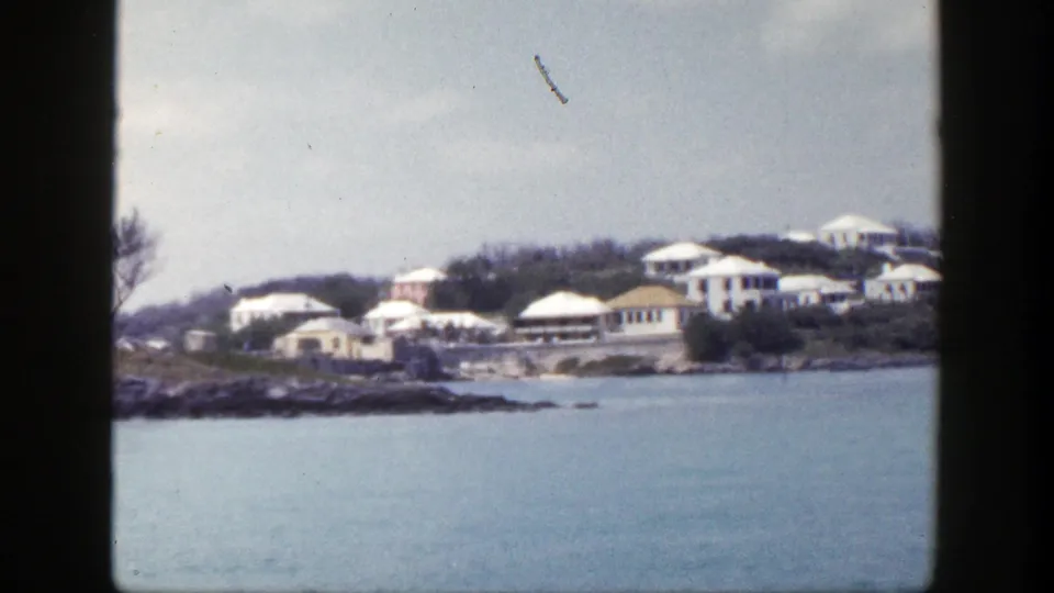 1948 New York coastal frame with low pale buildings, trees, rocky shoreline, and calm water in the foreground.