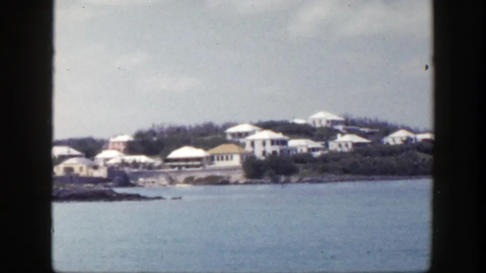 1948 New York home-movie still of white shoreline buildings, green vegetation, rocks, and calm blue water.