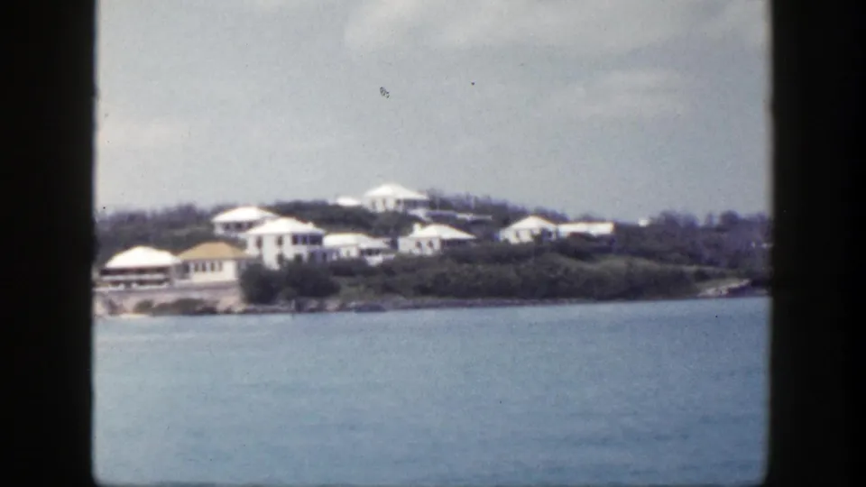 1948 coastal view from a moving boat, with white houses and trees along a low hillside across blue water.