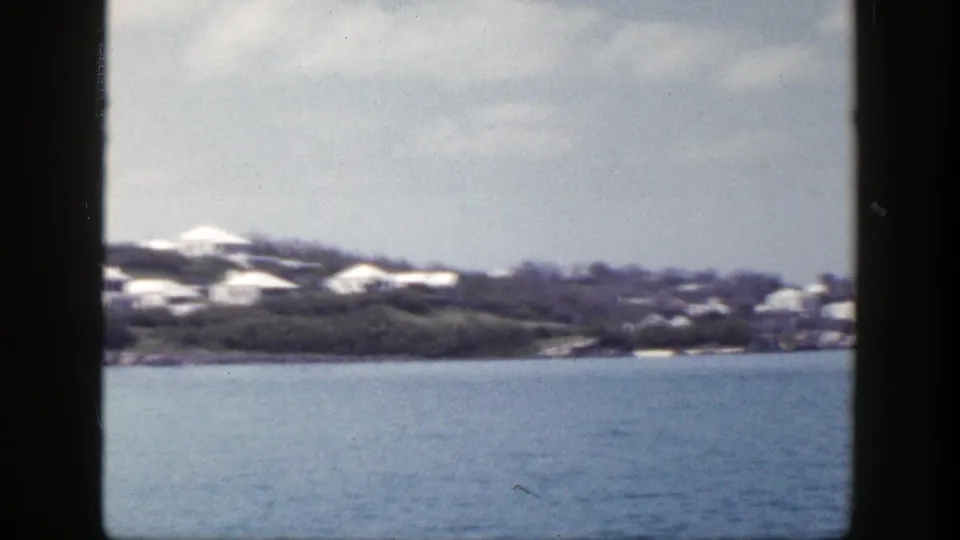 1948 8mm coastal frame with distant white houses on a wooded hillside and blue water in the foreground.