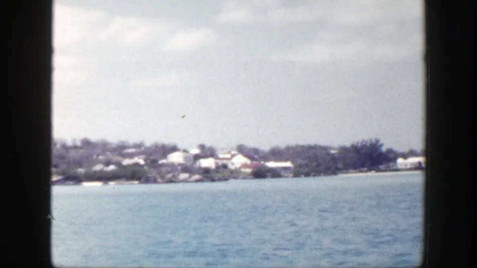 1948 New York 8mm frame showing a distant low shoreline, pale buildings, trees, and calm blue water through dark frame edges.
