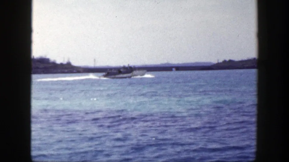 1948 New York 8mm frame with a small motorboat moving across blue water near a low shoreline and distant structures.