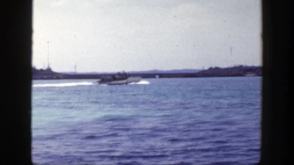 1948 New York 8mm frame of a small motorboat continuing across open water with a white wake and low land in the background.