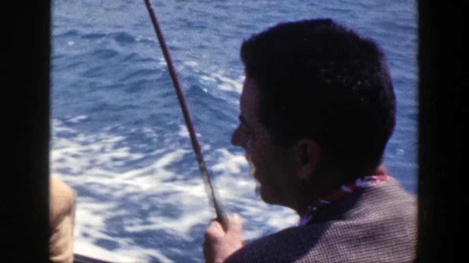 1948 New York home-movie frame of a man in profile holding a fishing rod while the boat moves through blue water.