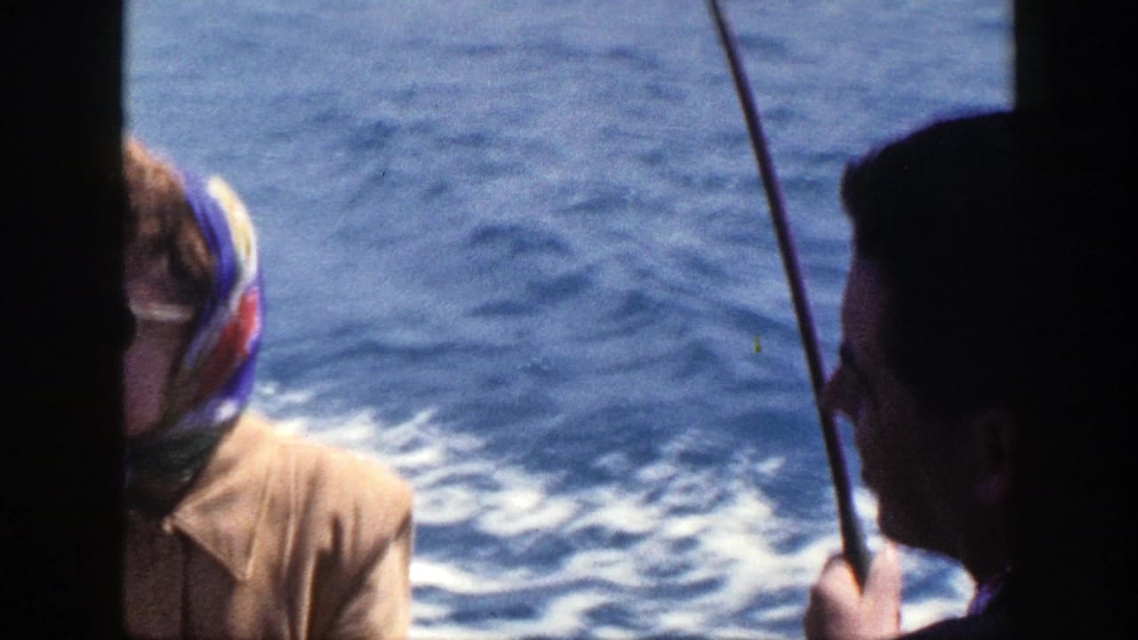 Man in profile and woman in headscarf sit near fishing rods with water behind them.