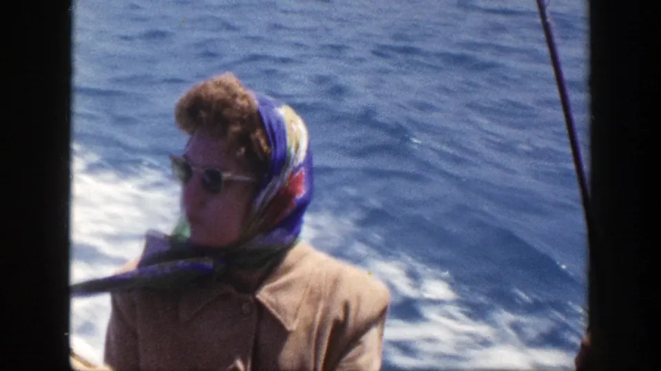 1948 New York home-movie frame showing a woman in sunglasses and a colorful headscarf on a moving boat, with wake and open water behind her.