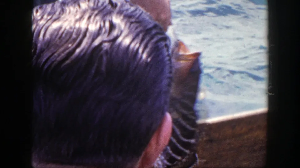 An adult seen from behind watches a fish held close to the boat in vintage 8mm New York fishing footage.