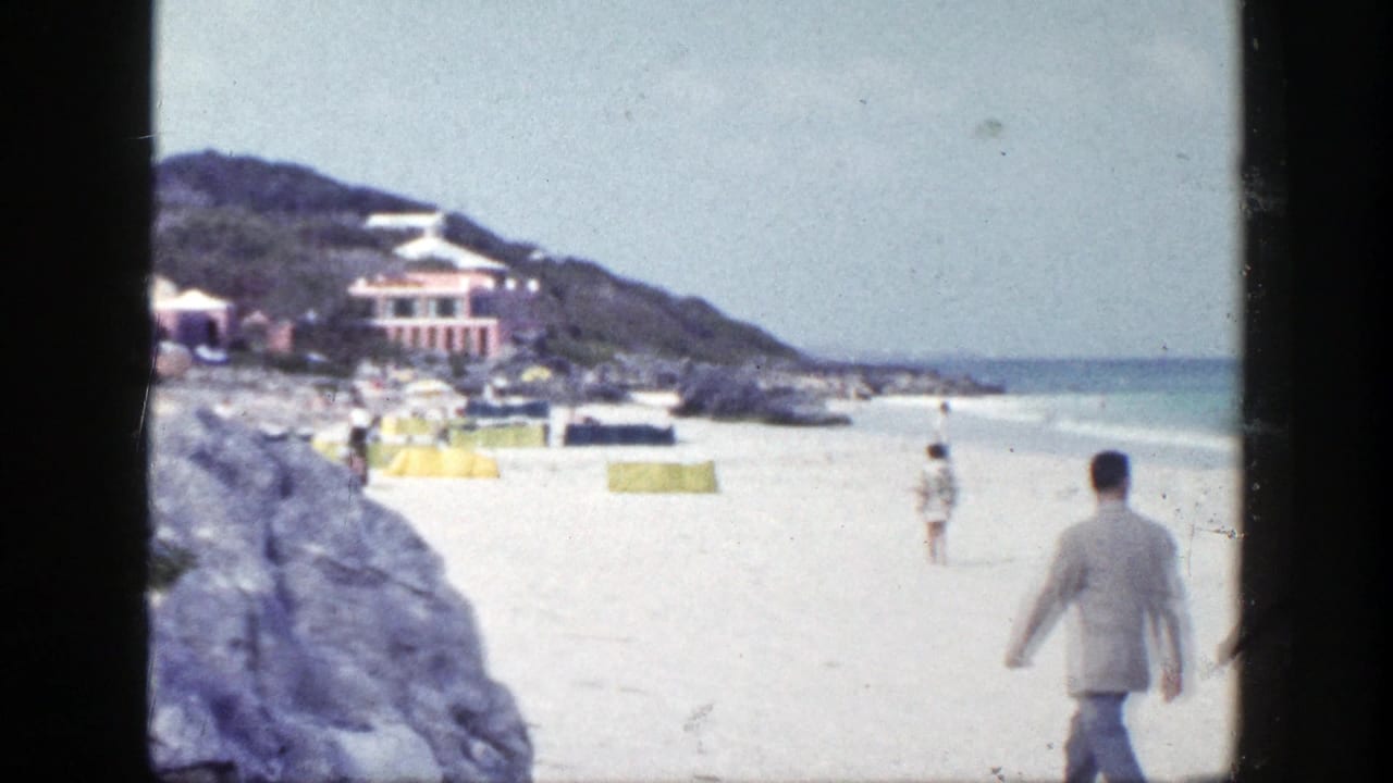 Man in a light suit walking along a sandy beach near rocks and ocean.