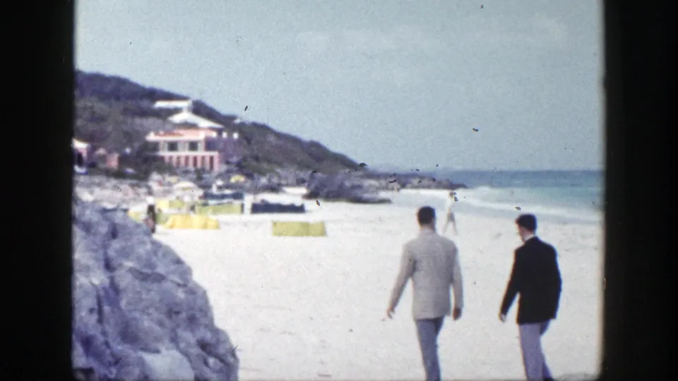 Two men in suits walk across a New York beach in 1948, with rocky shore, umbrellas, distant people, and ocean in view.