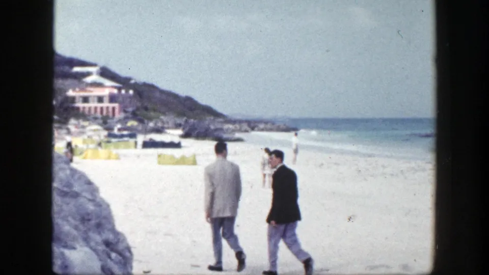 Two suited men walk and appear to talk on a 1948 New York beach, with surf, umbrellas, rocks, and hillside buildings behind them.