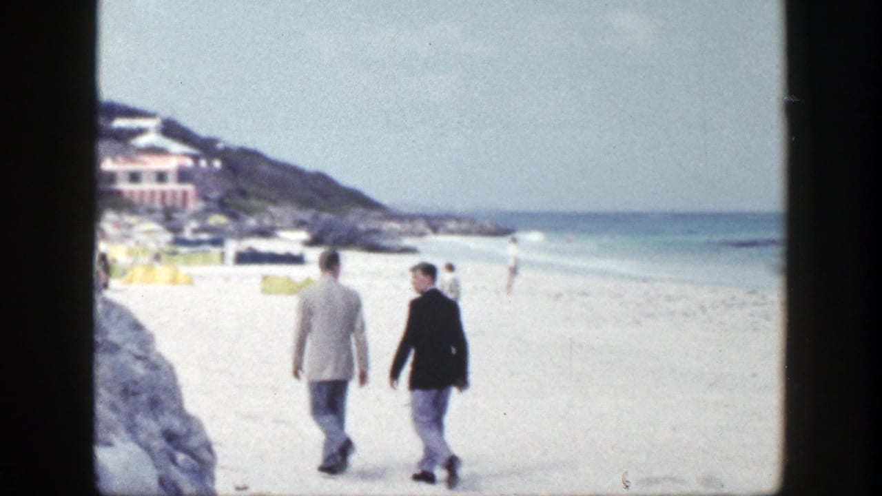 Two men in jackets walk away across a bright beach beside a rocky shoreline.