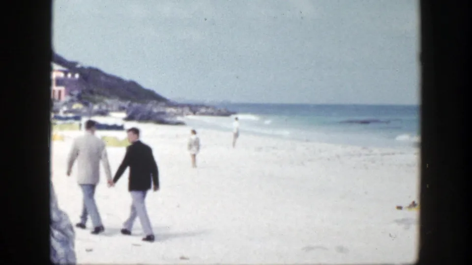 The two men continue along the New York beach, passing pale sand, low surf, distant swimmers, and hillside buildings in the 1948 home-movie scene.