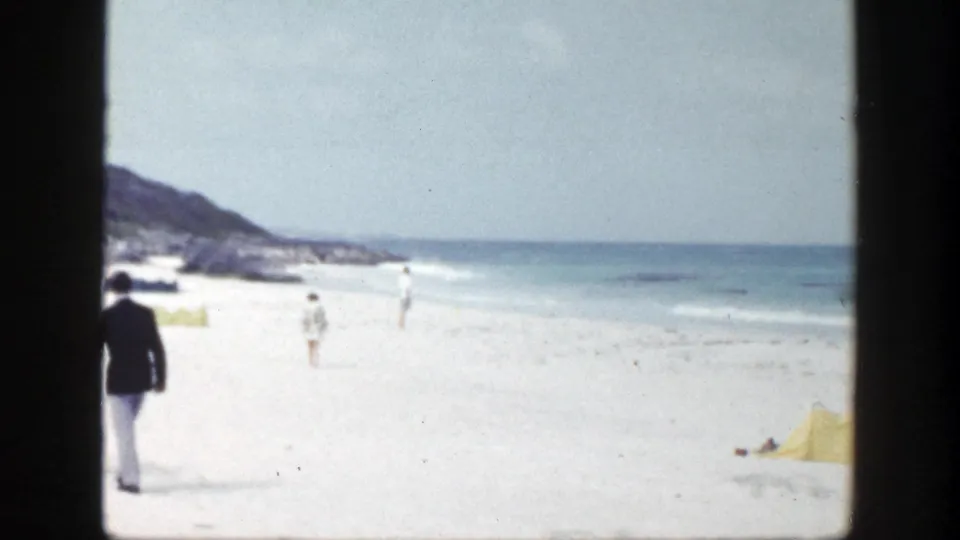 A suited figure moves out of frame on the left as the camera holds on the bright New York beach, surf, distant bathers, and shoreline structures.