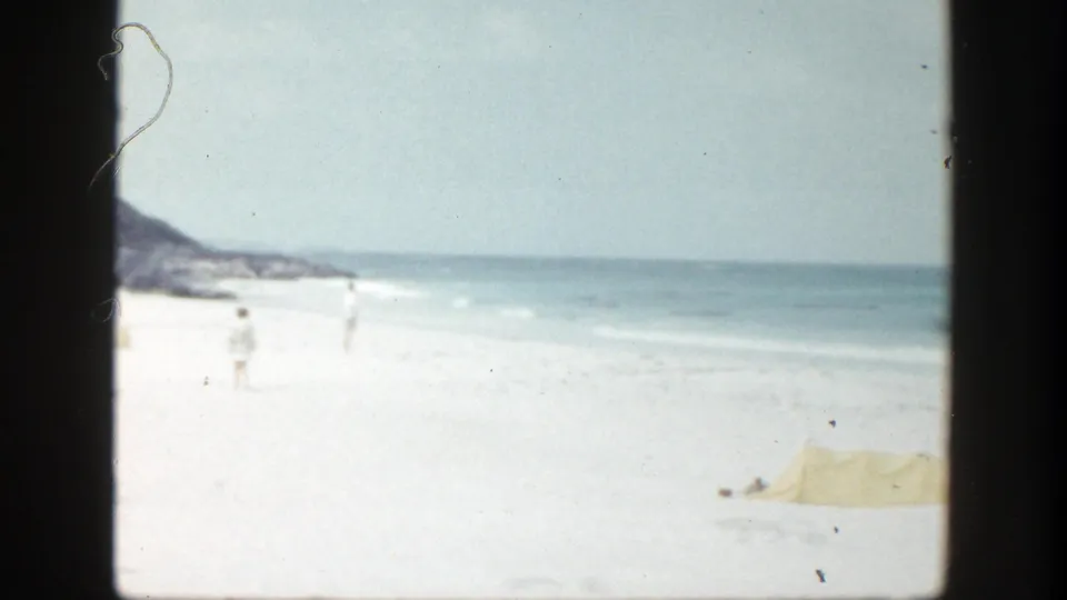 The final frame shows the New York shoreline after the men have passed, with pale sand, low surf, distant beachgoers, hillside coast, and yellow beach fabric.