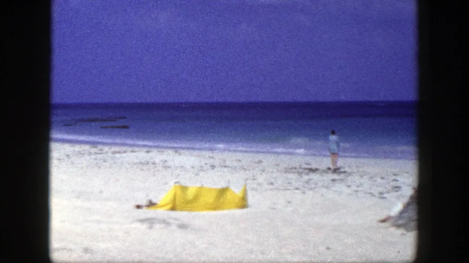A grainy 8mm view of a yellow beach tent on white sand, with a distant person standing near the New York shoreline.