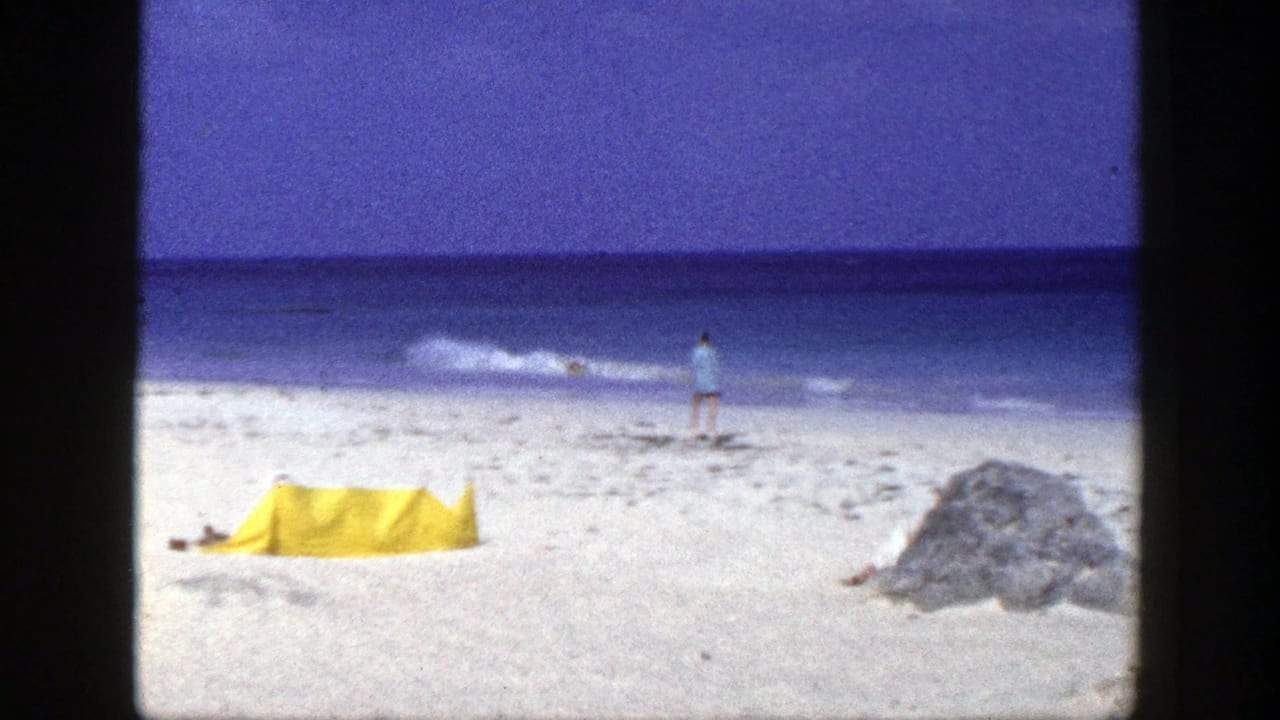 Man walking near the shoreline with a yellow tent and large rock on the sand.