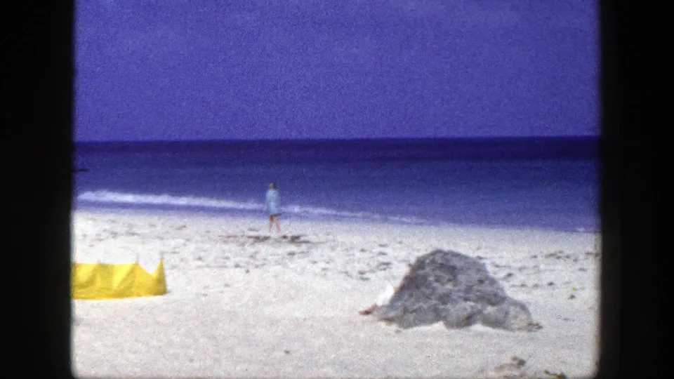 Home-movie beach view from 1948 metadata, showing a man near the waterline, yellow tent, large rock, white sand, and blue ocean.